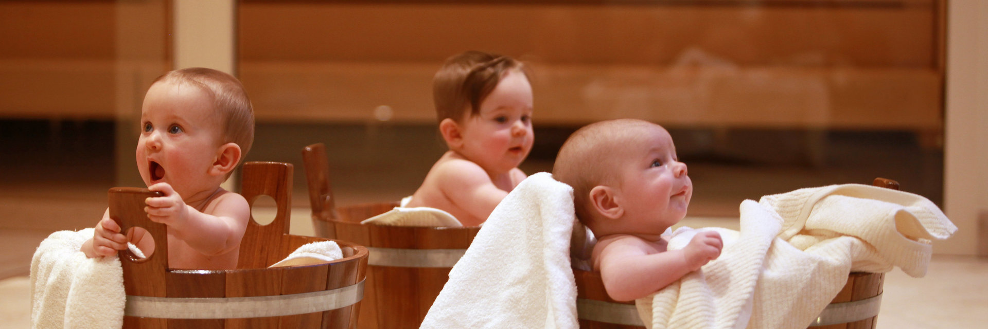 Drei Babys sitzen mit Handtüchern in Saunakübeln aus Holz.